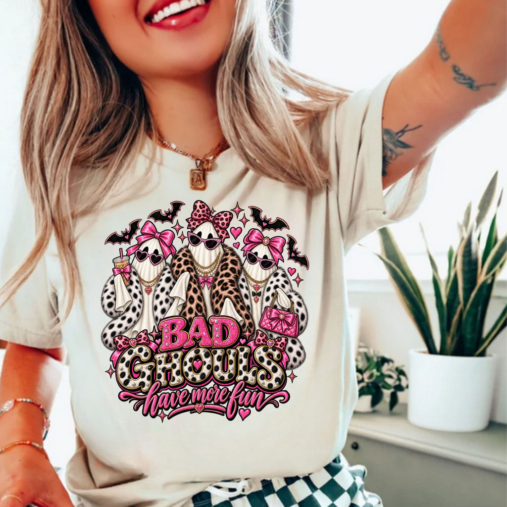 A woman wearing a cream-colored t-shirt with a graphic print of three ghosts dressed in faux fur outfits and pink bows, with the text 'BAD GHOULS have more fun' in pink and leopard print.