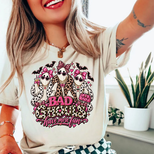 A woman wearing a cream-colored t-shirt with a graphic print of three ghosts dressed in faux fur outfits and pink bows, with the text 'BAD GHOULS have more fun' in pink and leopard print.