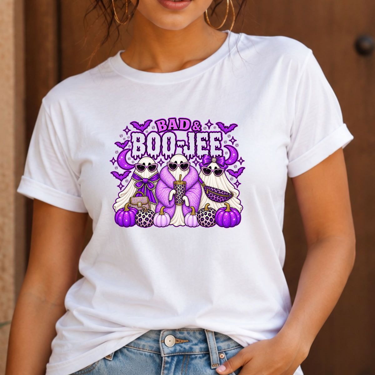 A woman wearing a white short-sleeve graphic T-shirt with a purple and pink Halloween design that reads 'BAD & BOO-JEE' above three cartoon ghosts and pumpkins, paired with blue jeans against a warm brown wooden background.
