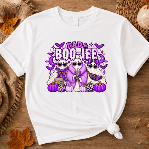 A white short-sleeve t-shirt with a graphic print of three cartoon ghosts wearing sunglasses and leopard-print clothing, standing in front of purple pumpkins and the text 'BAD BOO-JEE'.
