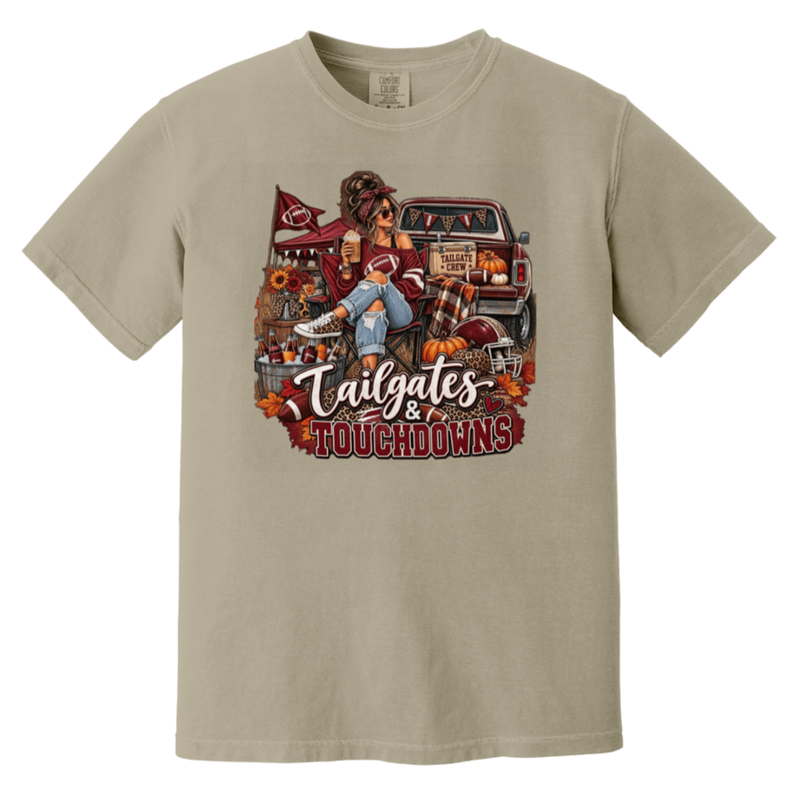 Beige graphic T-shirt with a detailed autumn football scene printed on the front, including a truck, pumpkins, sunflowers, and the text 'Tailgates & Touchdowns'.