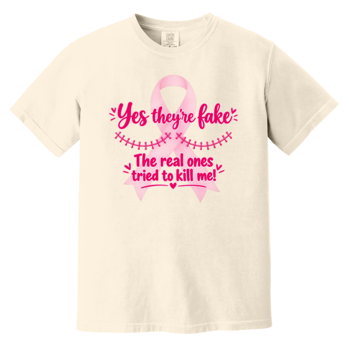Beige short-sleeve t-shirt with a pink graphic on the front. The graphic features a pink ribbon and the text 'Yes they're fake' and 'The real ones tried to kill me!' in pink lettering.