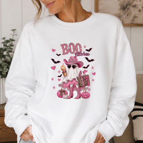 A woman wearing a white crewneck sweatshirt with a pink and black 'Boo Bitches' Halloween graphic featuring a ghost, cowboy hat, sunglasses, and pumpkins.