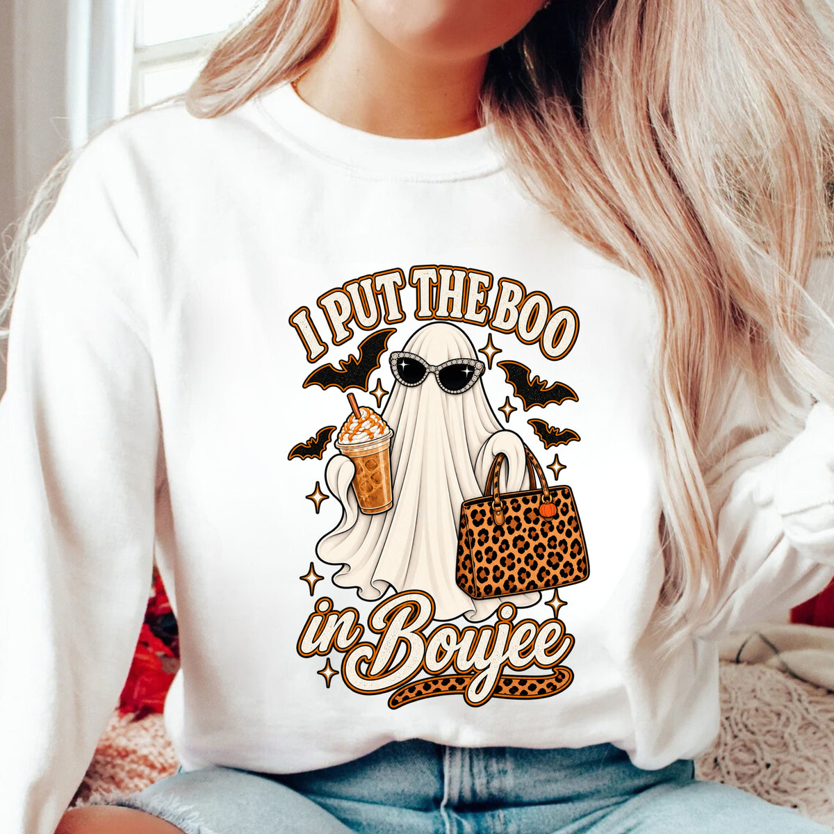 A woman wearing a white crewneck sweatshirt with a graphic print that reads 'I PUT THE BOO in Boujee' featuring a ghost in sunglasses holding a drink and a leopard-print handbag.