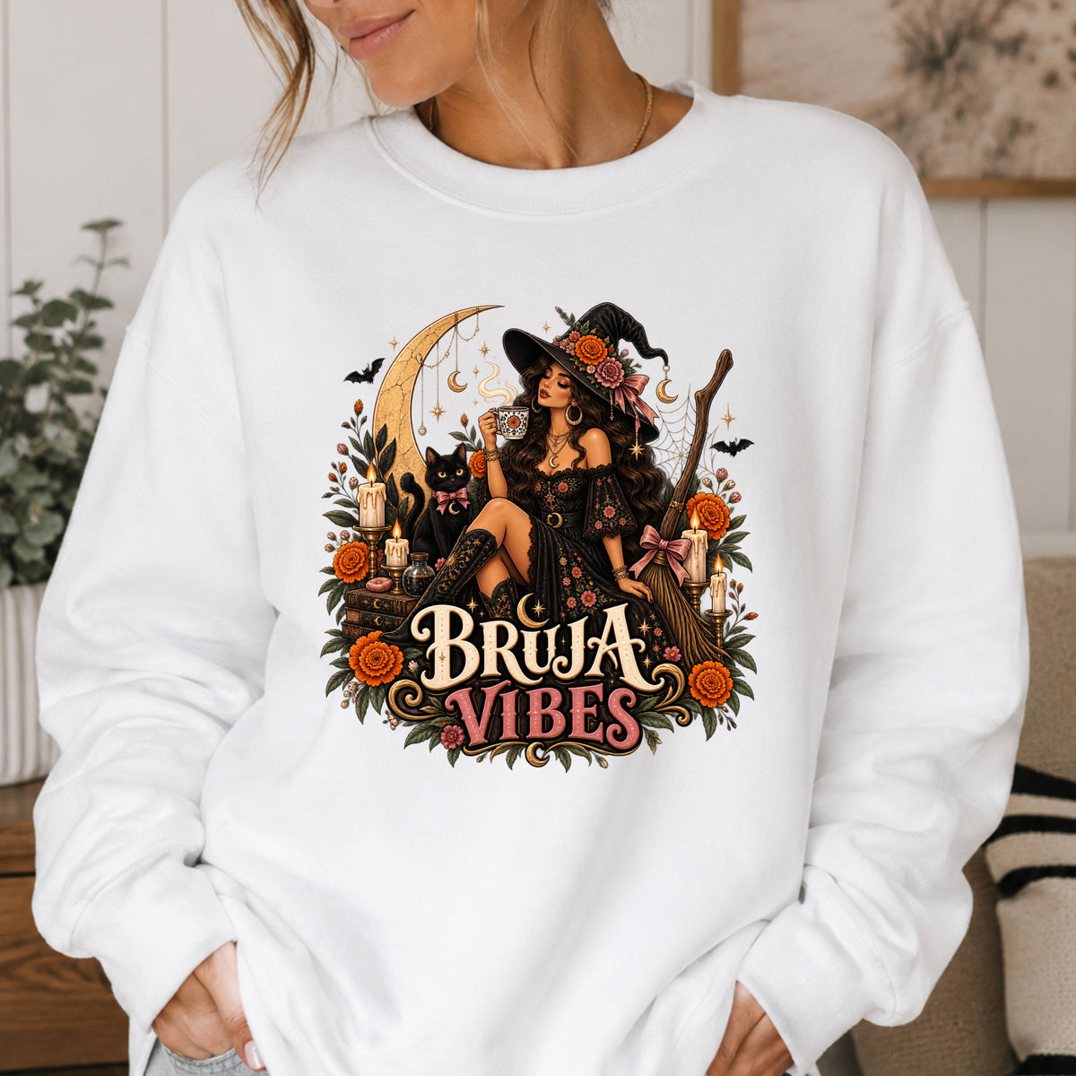 A woman wearing a white crewneck sweatshirt with a large graphic print on the front. The graphic features a witch in a black hat sitting on a broomstick next to a black cat, surrounded by orange flowers and the text 'BRUJA VIBES'.
