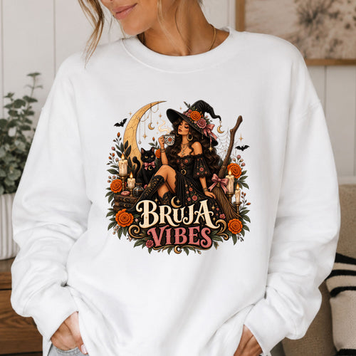 A woman wearing a white crewneck sweatshirt with a large graphic print on the front. The graphic features a witch in a black hat sitting on a broomstick next to a black cat, surrounded by orange flowers and the text 'BRUJA VIBES'.