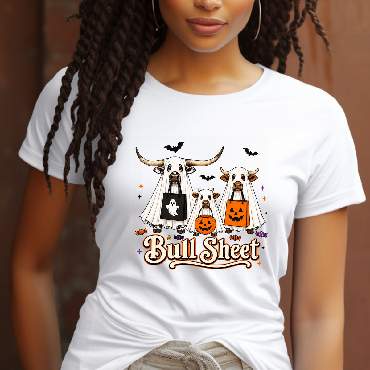 A woman wearing a white graphic t-shirt with a cartoon design of three cows dressed in Halloween costumes and the text 'Bull Sheet'.