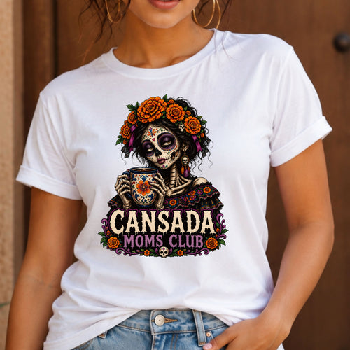 A woman wearing a white graphic t-shirt featuring a Day of the Dead sugar skull design with orange flowers and the text 'CANSADA MOMS CLUB'.