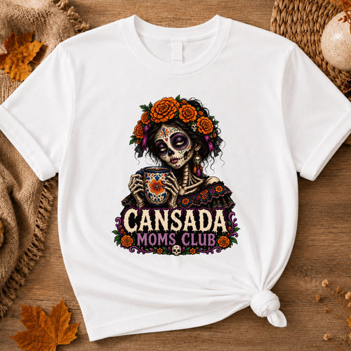 A white short-sleeve t-shirt with a colorful Day of the Dead sugar skull graphic and the text 'CANSADA MOMS CLUB'. The t-shirt is styled in a flat-lay on a rustic wooden background with autumn props like a burlap sack, a woven placemat, and dried leaves.