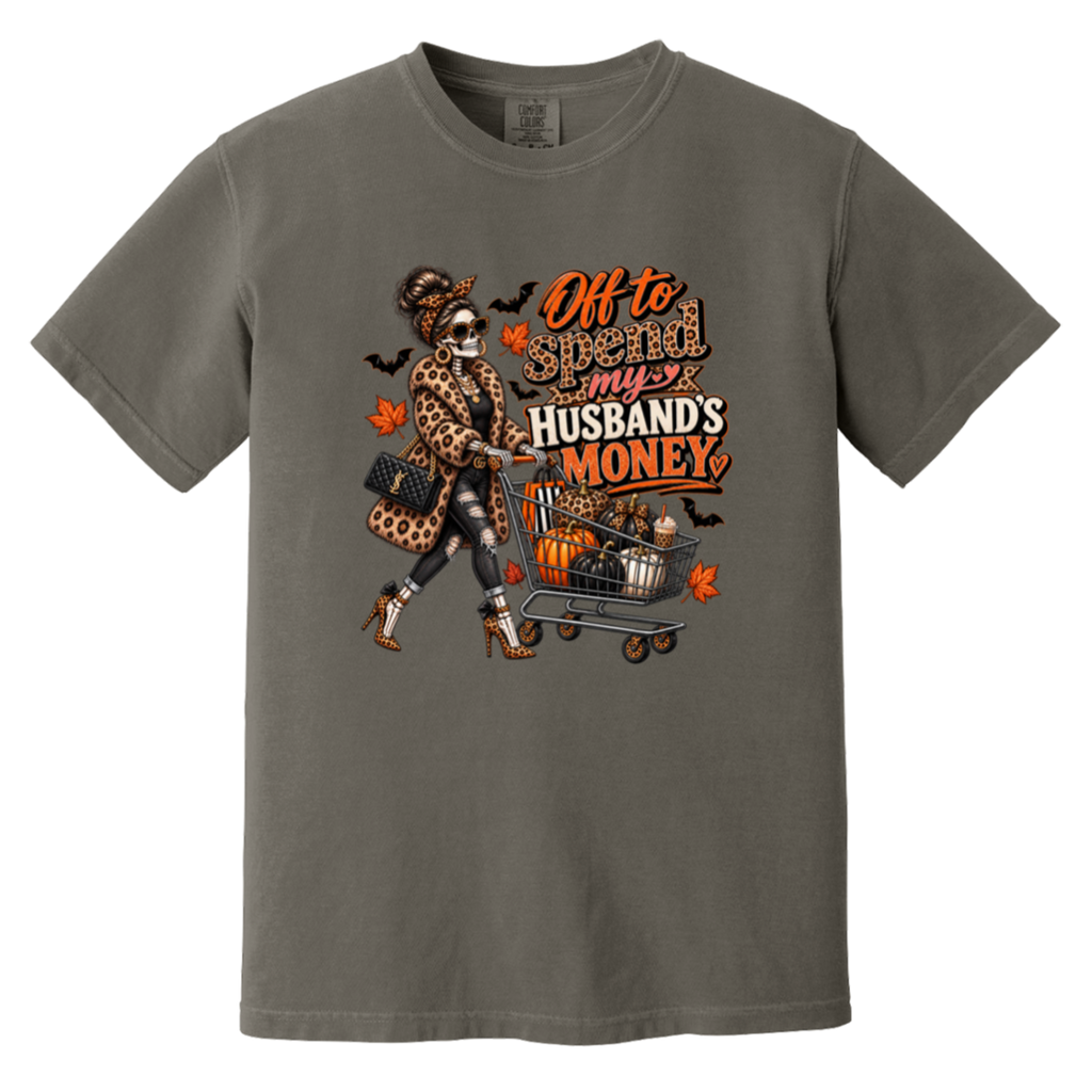 A dark gray tshirt featuring a skeleton wearing a leopard print fur coat, pushing a shopping cart full of pumpkins with the text ‘off to spend my husbands money’ 