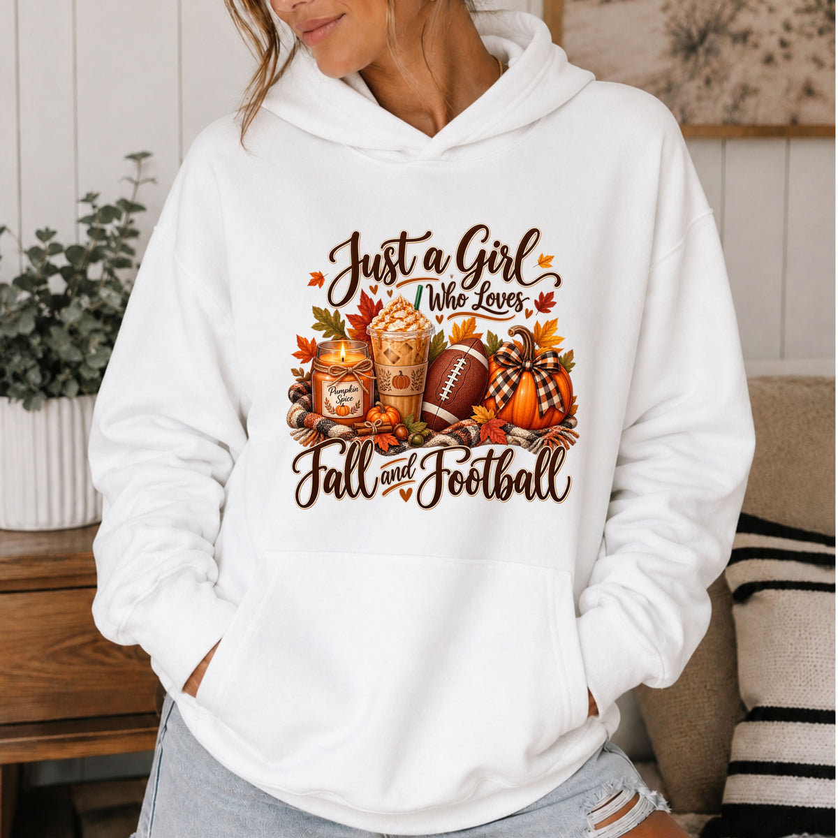 A woman wearing a white pullover hoodie with a 'Just a Girl Who Loves Fall and Football' graphic featuring autumn leaves, a football, and a pumpkin. The photo is taken in a cozy indoor setting.