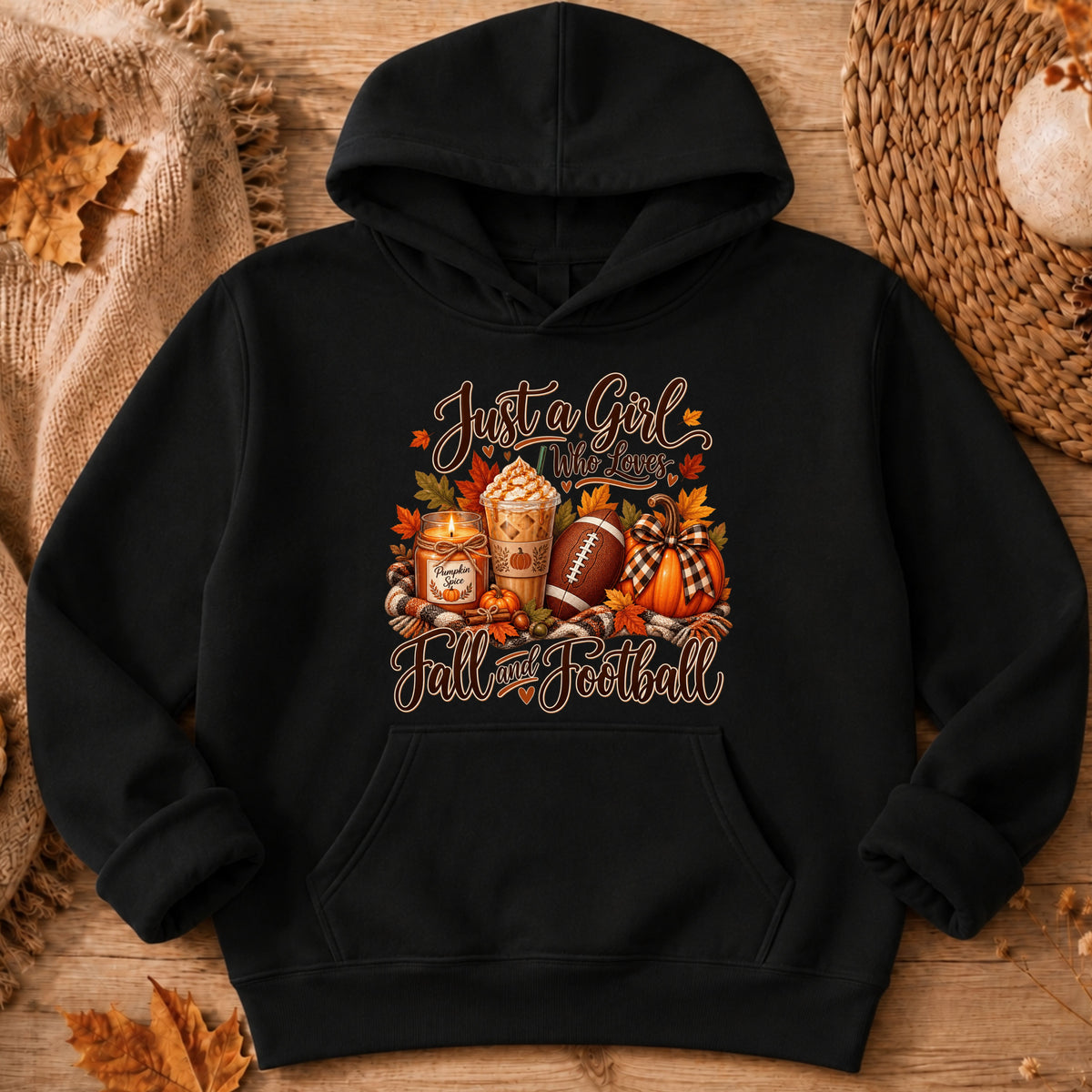 A black pullover hoodie laid flat on a rustic wood surface with autumn-themed props. The front has a large fall football graphic with the phrase 'Just a Girl Who Loves Fall Football' in warm orange-brown lettering, including footballs, pumpkins, leaves, and a hot dog cup.