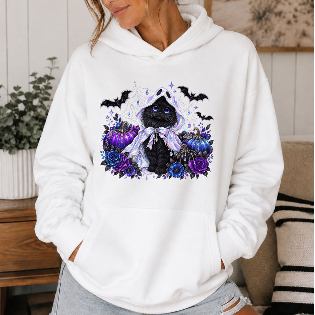 A woman wearing a white pullover hoodie with a Halloween-themed graphic print of a black cat, purple pumpkins, blue roses, bats, and spiderwebs on the chest, set in a cozy indoor environment.