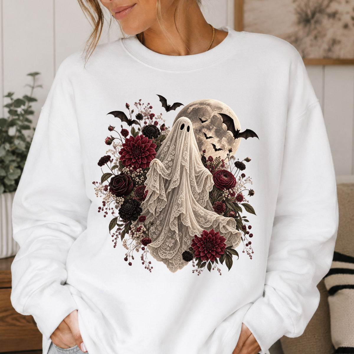 A woman wearing a white crewneck sweatshirt with a gothic graphic of a ghost, dark red flowers, bats, and a full moon.