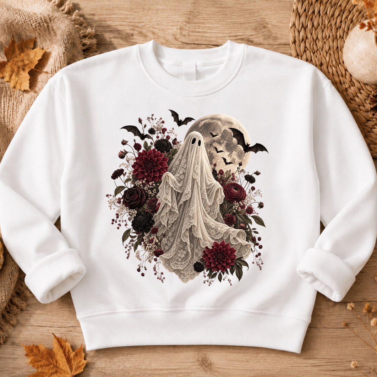 A white crewneck sweatshirt laid flat on a rustic wooden surface, featuring a gothic-style graphic of a ghostly figure draped in lace, surrounded by dark red and black flowers, bats, and a full moon.