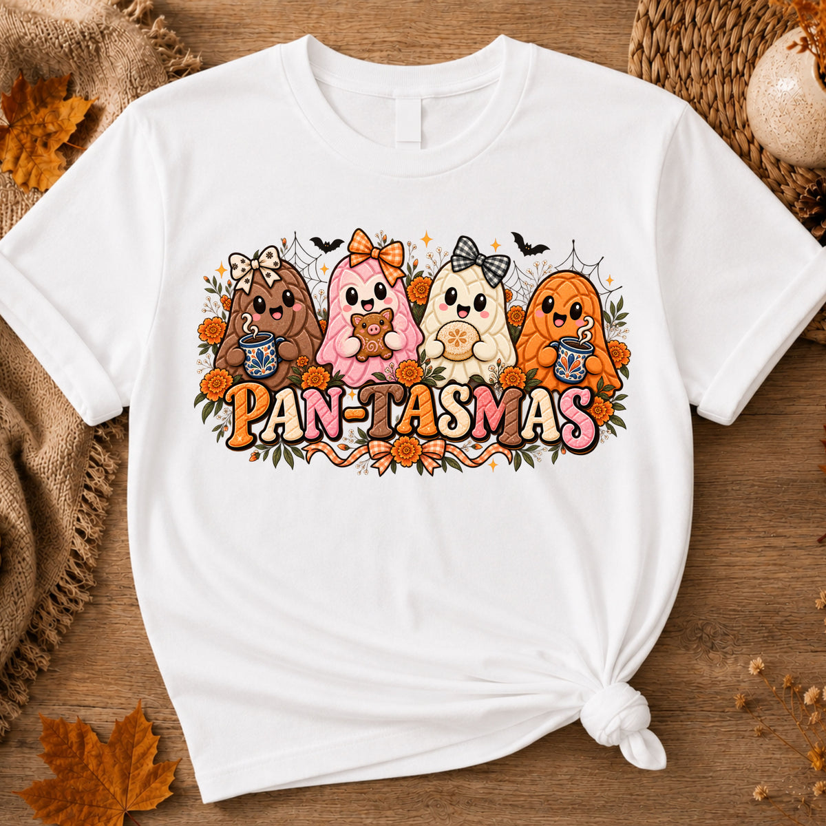 A white short-sleeve graphic T-shirt laid flat on a rustic wood surface with autumn props. The shirt has a colorful front print showing four cute ghost-like sweet bread characters and the text 'PAN-TASMAS.