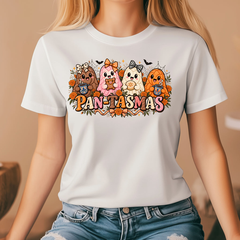 A woman wearing a white short-sleeve graphic T-shirt with a colorful cartoon design of four ghost shaped pan dulce above the text 'PAN-TASMAS', paired with blue jeans in a softly lit indoor setting.