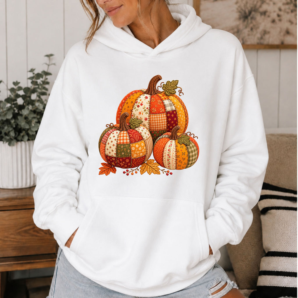 A woman wearing a white pullover hoodie with a colorful patchwork pumpkin graphic on the front, standing in a cozy indoor setting.