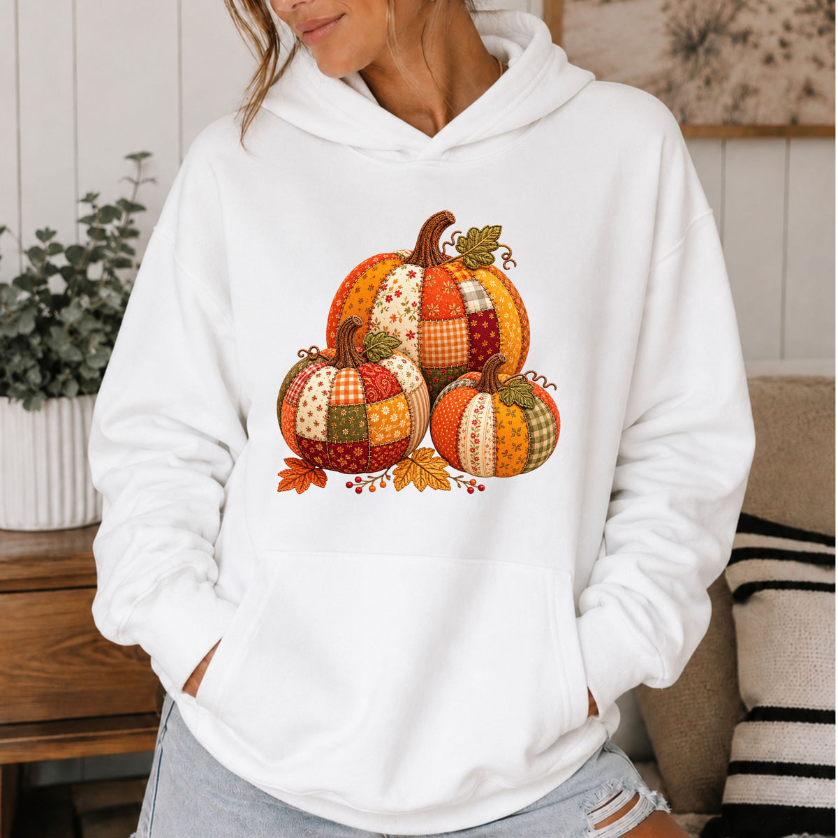 A woman wearing a white pullover hoodie with a colorful patchwork pumpkin graphic on the front, standing in a cozy indoor setting.