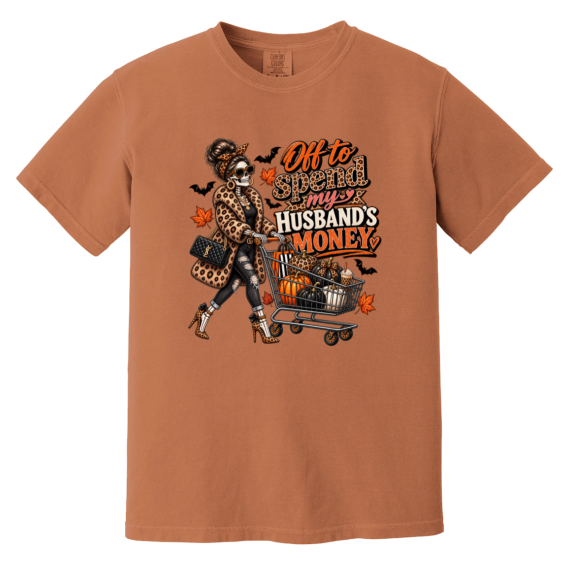 A rust orange tshirt featuring a skeleton wearing a leopard print fur coat, pushing a shopping cart full of pumpkins with the text ‘off to spend my husbands money’ 