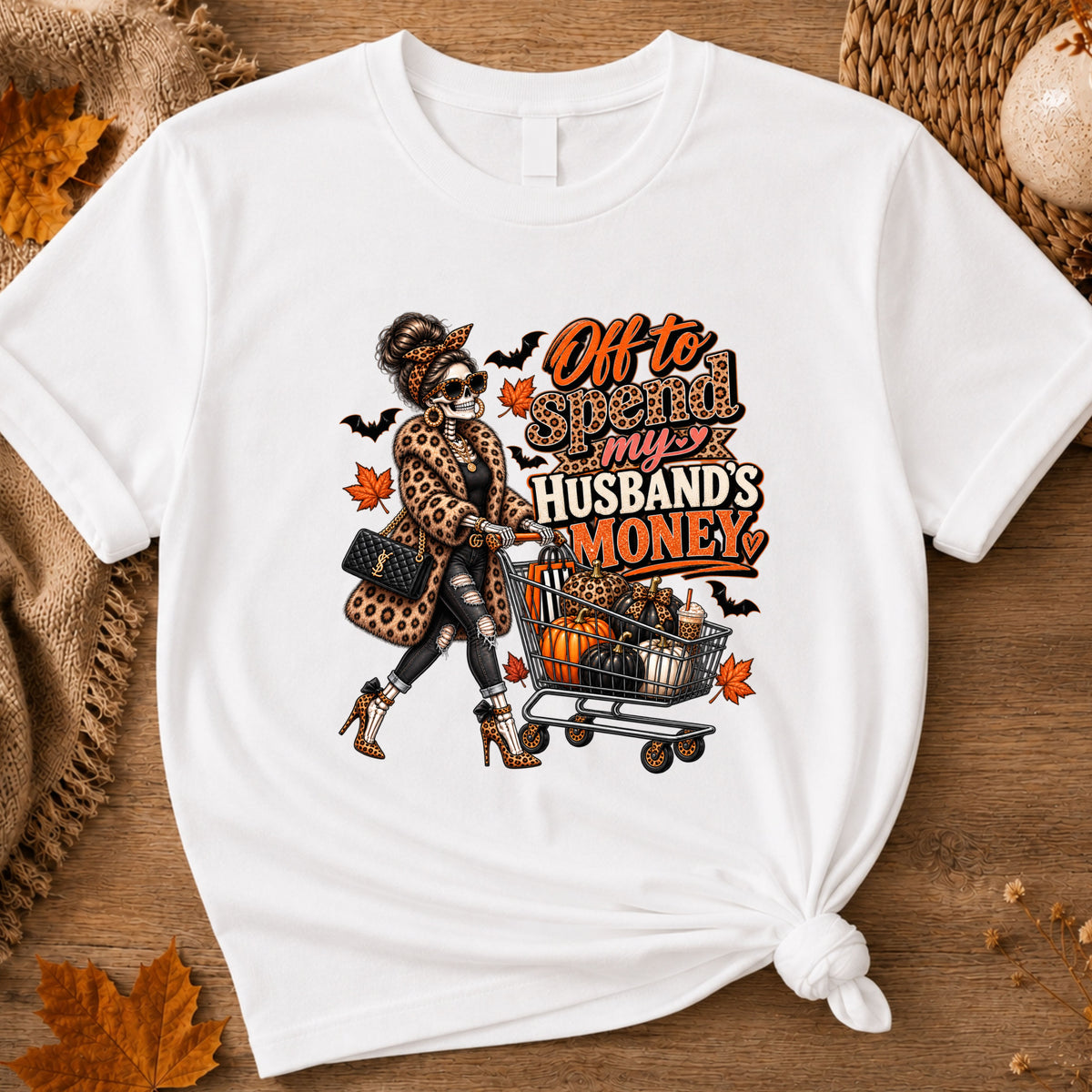 A white tshirt featuring a skeleton wearing a leopard print fur coat, pushing a shopping cart full of pumpkins with the text ‘off to spend my husbands money’ 