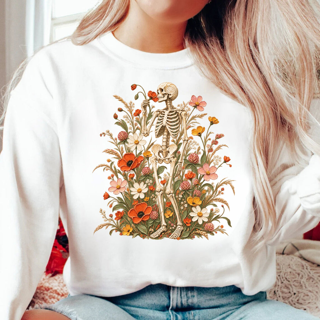 A woman wearing a white crewneck sweatshirt with a detailed graphic of a skeleton surrounded by colorful wildflowers. The sweatshirt is shown in a cozy indoor setting with light blue jeans.