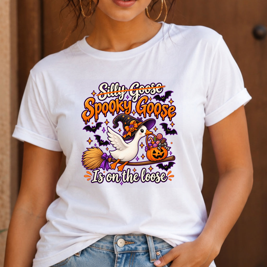 A woman wearing a white short-sleeve graphic T-shirt with a Halloween design featuring the text 'Silly Goose Spooky Goose Is on the loose' and an illustration of a white goose, witch hat, broom, pumpkin, and bats.