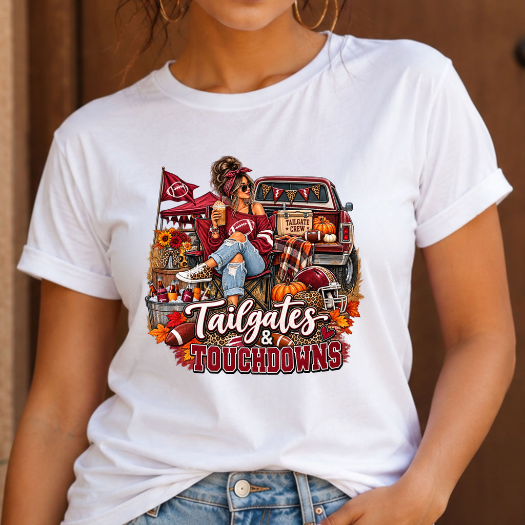 A woman wearing a white short-sleeve graphic T-shirt with a detailed autumn football scene printed on the front, including a truck, pumpkins, sunflowers, and the text 'Tailgates & Touchdowns'.