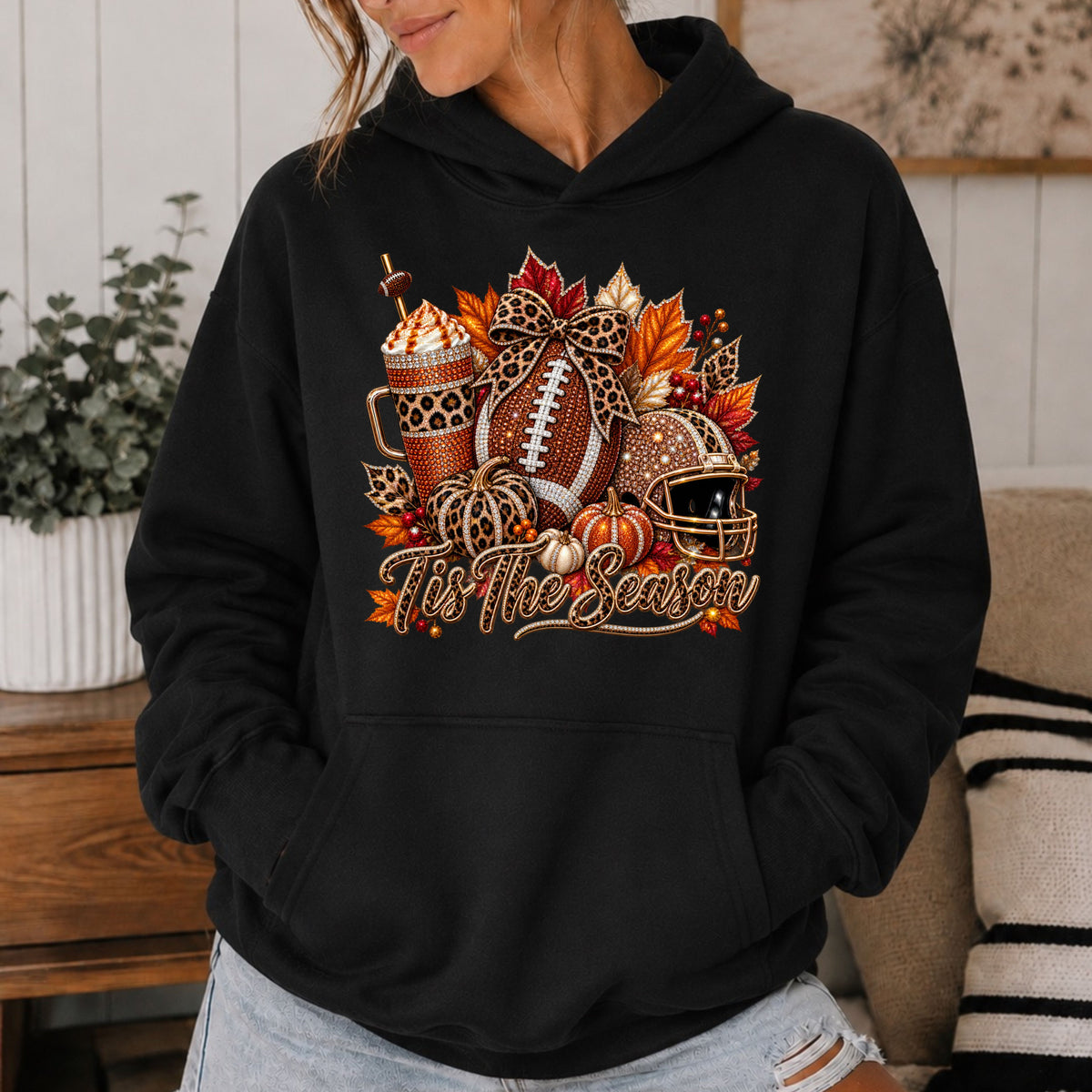 A black hoodie with a fall themed graphic featuring a football, iced coffee, football helmet, and a text saying tis the season