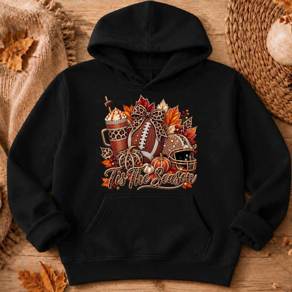 A black pullover hoodie laid flat on a rustic wood surface with autumn props. The front has a large seasonal graphic featuring a football, iced coffee,  pumpkin, leaves, and the phrase “tis The Season,” in warm fall colors.
