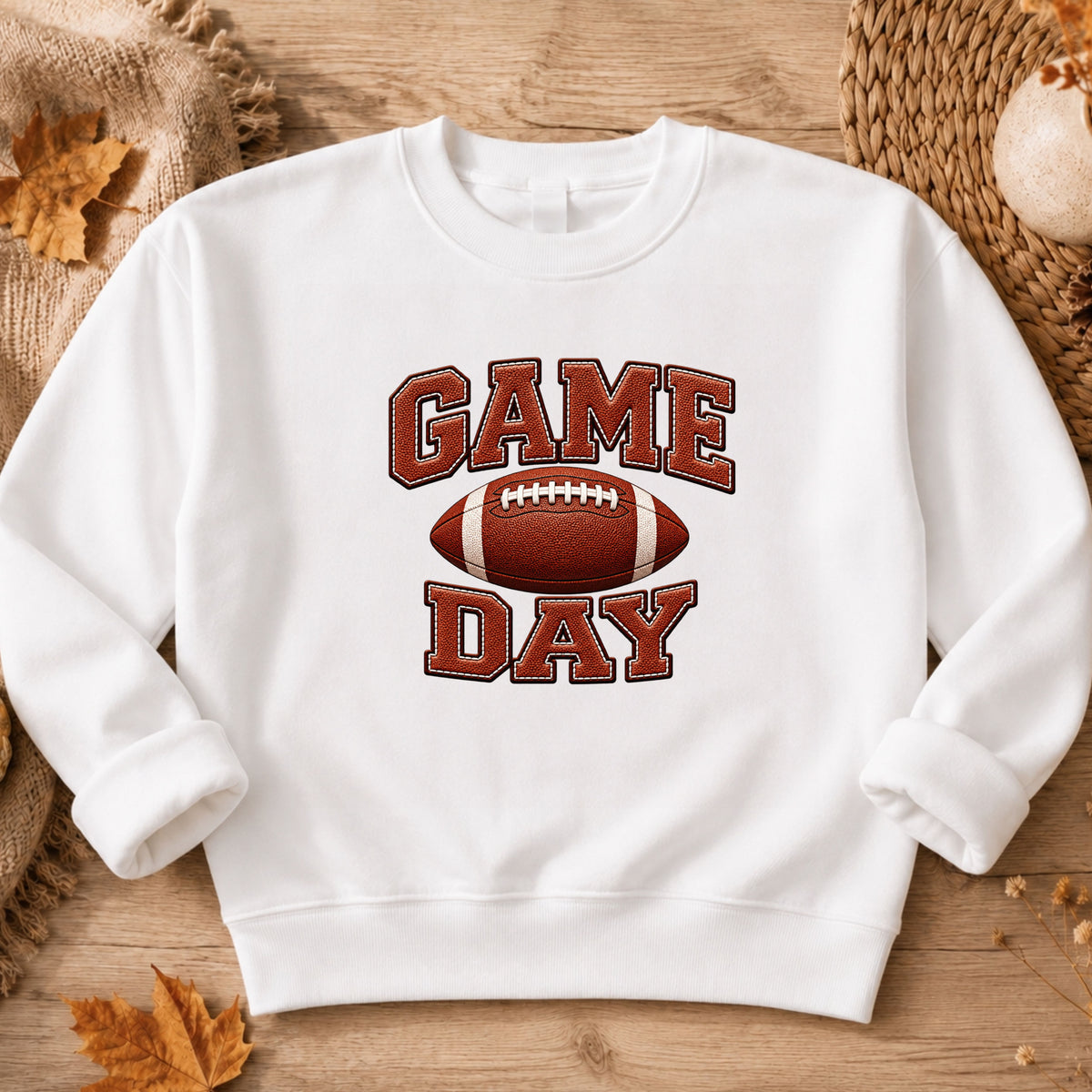 A white crewneck sweatshirt featuring a 'GAME DAY' football graphic, laid flat on a rustic wooden surface with autumn leaves and woven textures.