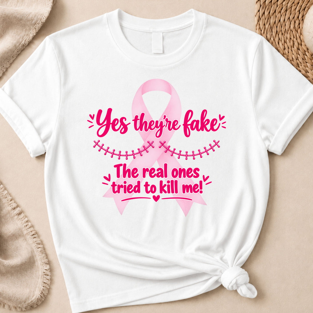 White short-sleeve T-shirt laid flat with a pink awareness ribbon graphic and the text 'Yes they're fake. The real ones tried to kill me!' in bright pink, styled on a beige surface with soft fabric and woven mat props.
