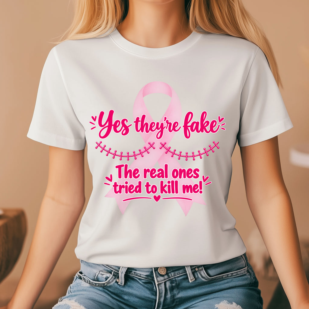 A woman wearing a white short-sleeve graphic T-shirt with a pink ribbon and the text 'Yes they're fake. The real ones tried to kill me!' in pink lettering, paired with blue jeans in a softly lit indoor setting.