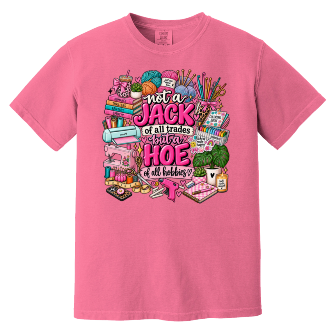 A pink short-sleeve crewneck t-shirt featuring a colorful graphic print on the front. The graphic contains various hand-drawn sewing and crafting tools (spools of yarn, a sewing machine, scissors, fabric) surrounding the text 'not a JACK of all trades but a HOE of all hobbies' in stylized pink and white lettering.
