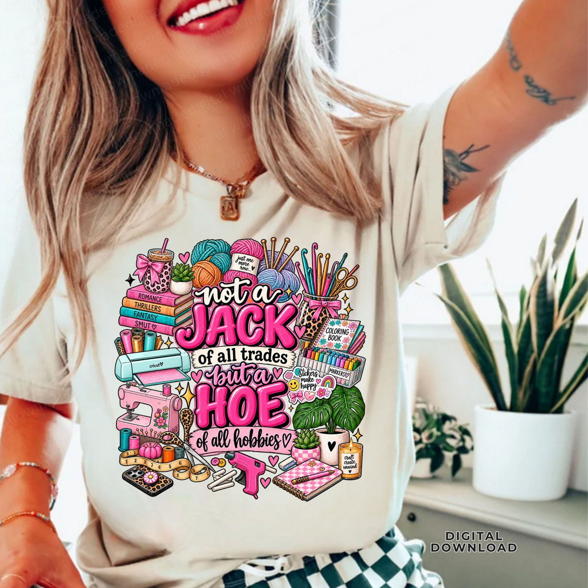 A woman wearing a cream-colored graphic t-shirt with a colorful design that reads 'not a JACK of all trades but a HOE of all hobbies' surrounded by sewing and crafting items. The photo is taken in a bright indoor setting with a snake plant in the background.