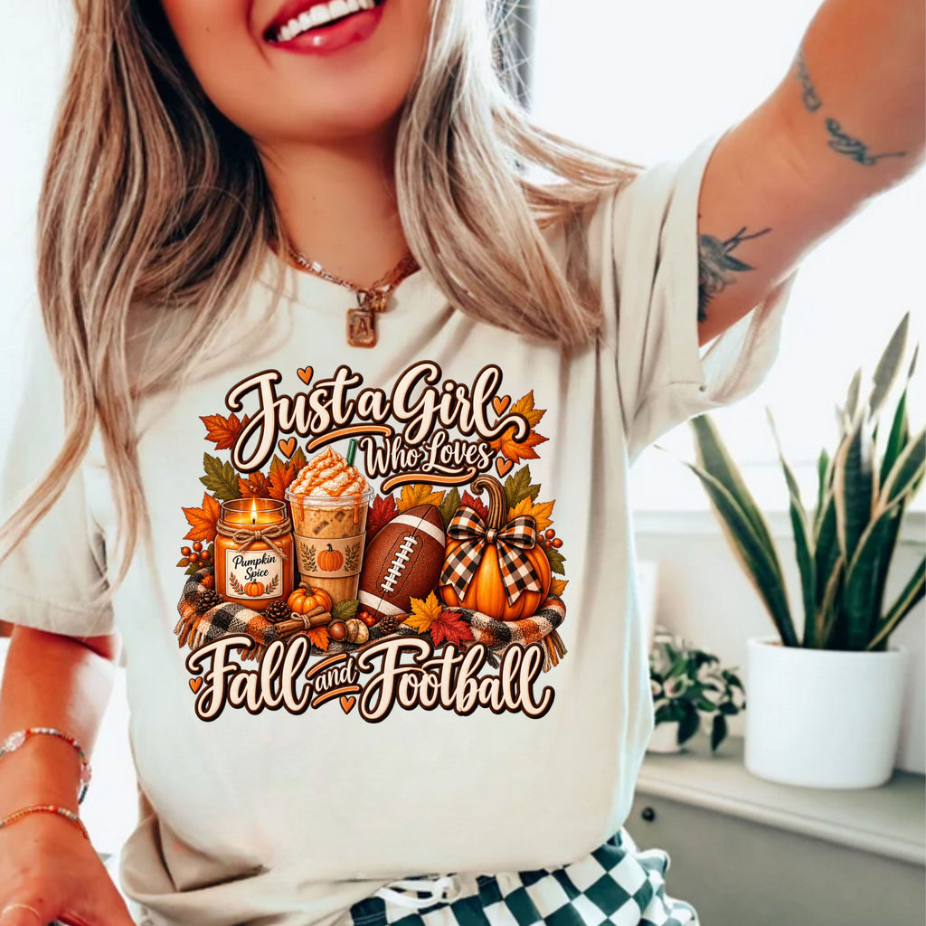 A woman wearing a cream-colored t-shirt with a colorful autumn graphic that reads 'Just a Girl Who Loves Fall and Football' with pumpkins, a football, a fall candle and iced coffee.