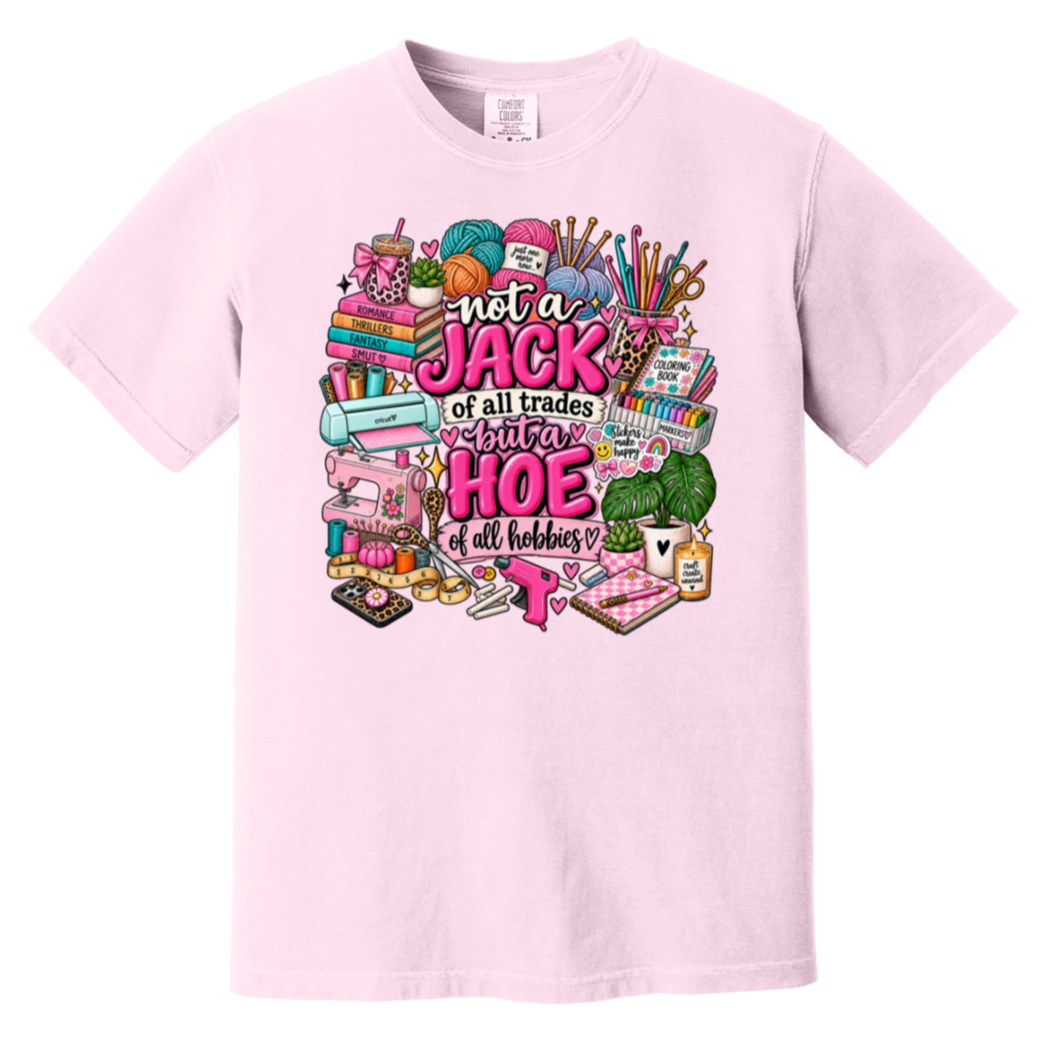 A light pink short-sleeve crewneck t-shirt featuring a colorful graphic print on the front. The graphic contains various sewing and crafting tools (spools of thread, a sewing machine, knitting needles, a green yardage roll) surrounding the text 'not a JACK of all trades but a HOE of all hobbies' in pink and white lettering.