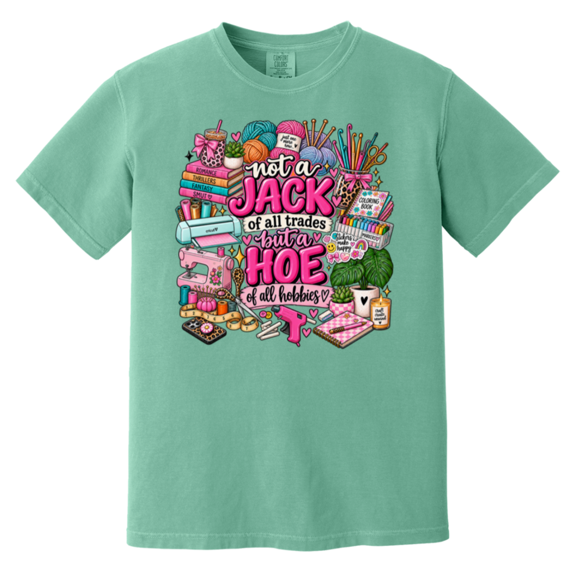 A mint green short-sleeve t-shirt with a colorful graphic print on the front. The graphic features various sewing and crafting tools (spools of thread, a sewing machine, knitting needles, fabric) surrounding the text 'not a JACK of all trades but a HOE of all hobbies' in pink and white lettering.