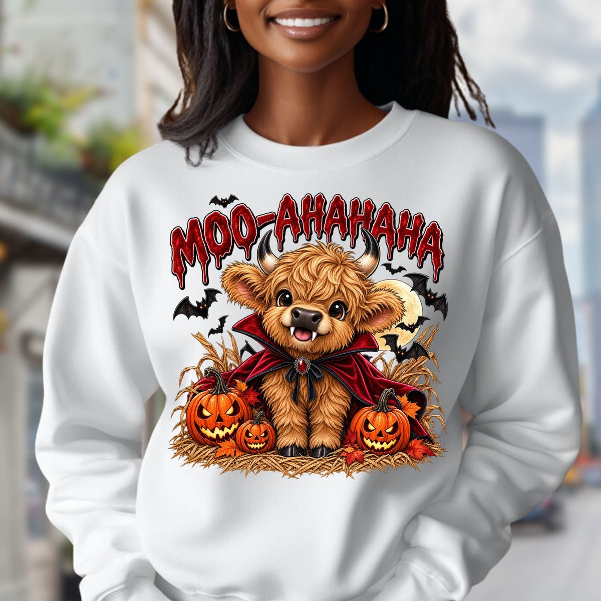 white crewneck sweatshirt featuring a cartoon graphic of a fluffy brown Scottish highland cow dressed in a red cape, surrounded by jack-o'-lanterns, with the text 'MOO-AAHAHAHA' in red above it.
